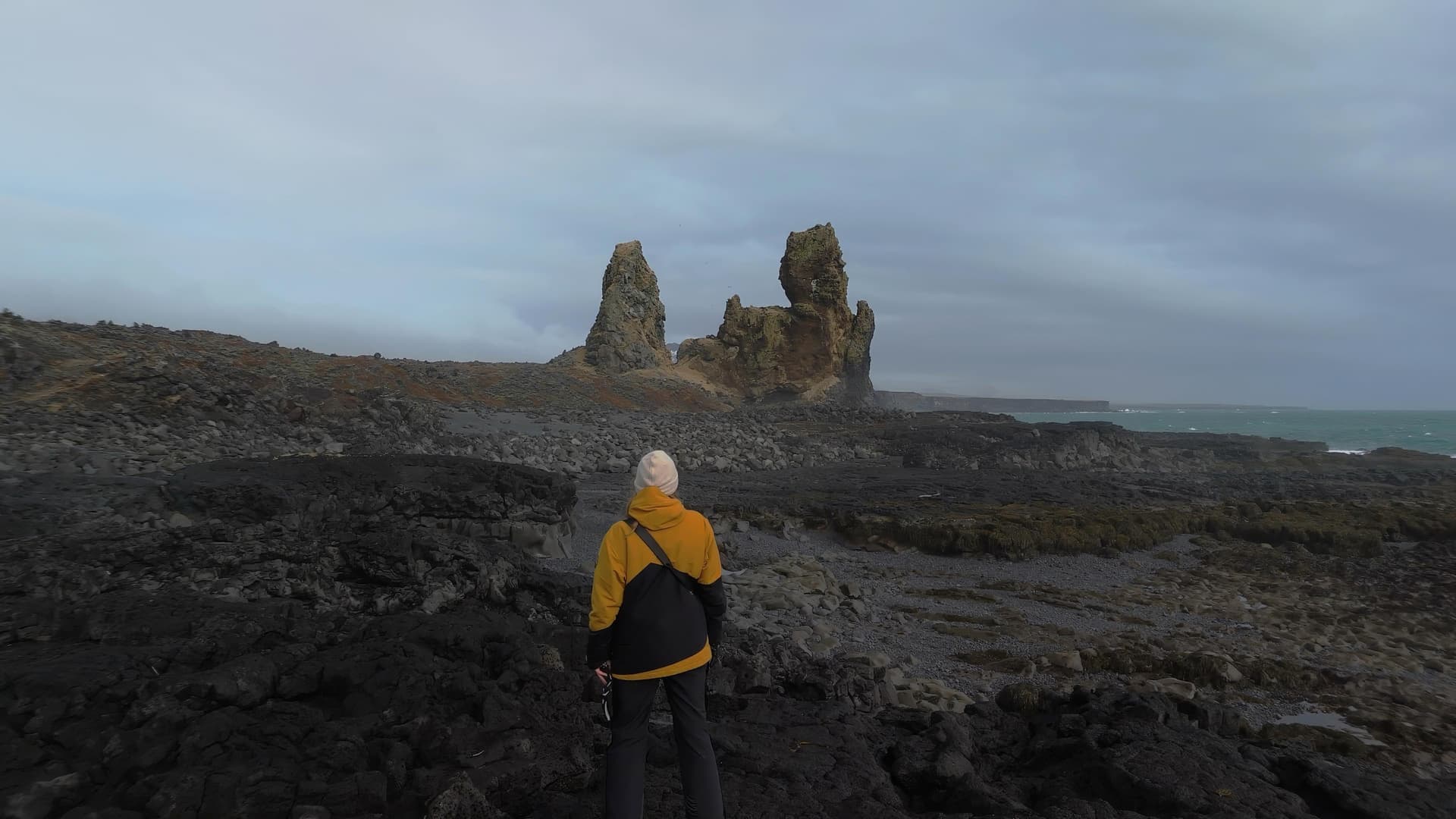 A lone figure on a black lava shore, facing weathered sea stacks under a heavy sky